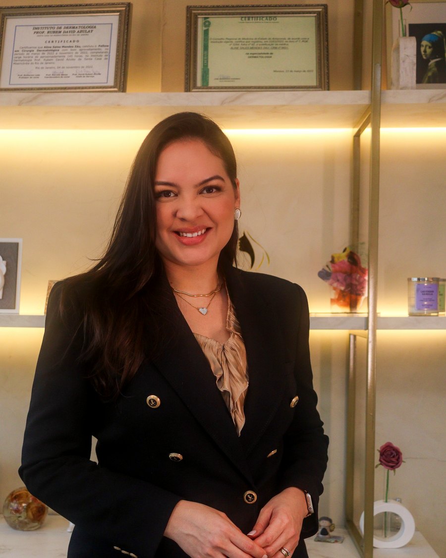 Dra. Aline Záu em pé no consultório, diante da parede com os certificados de formação.