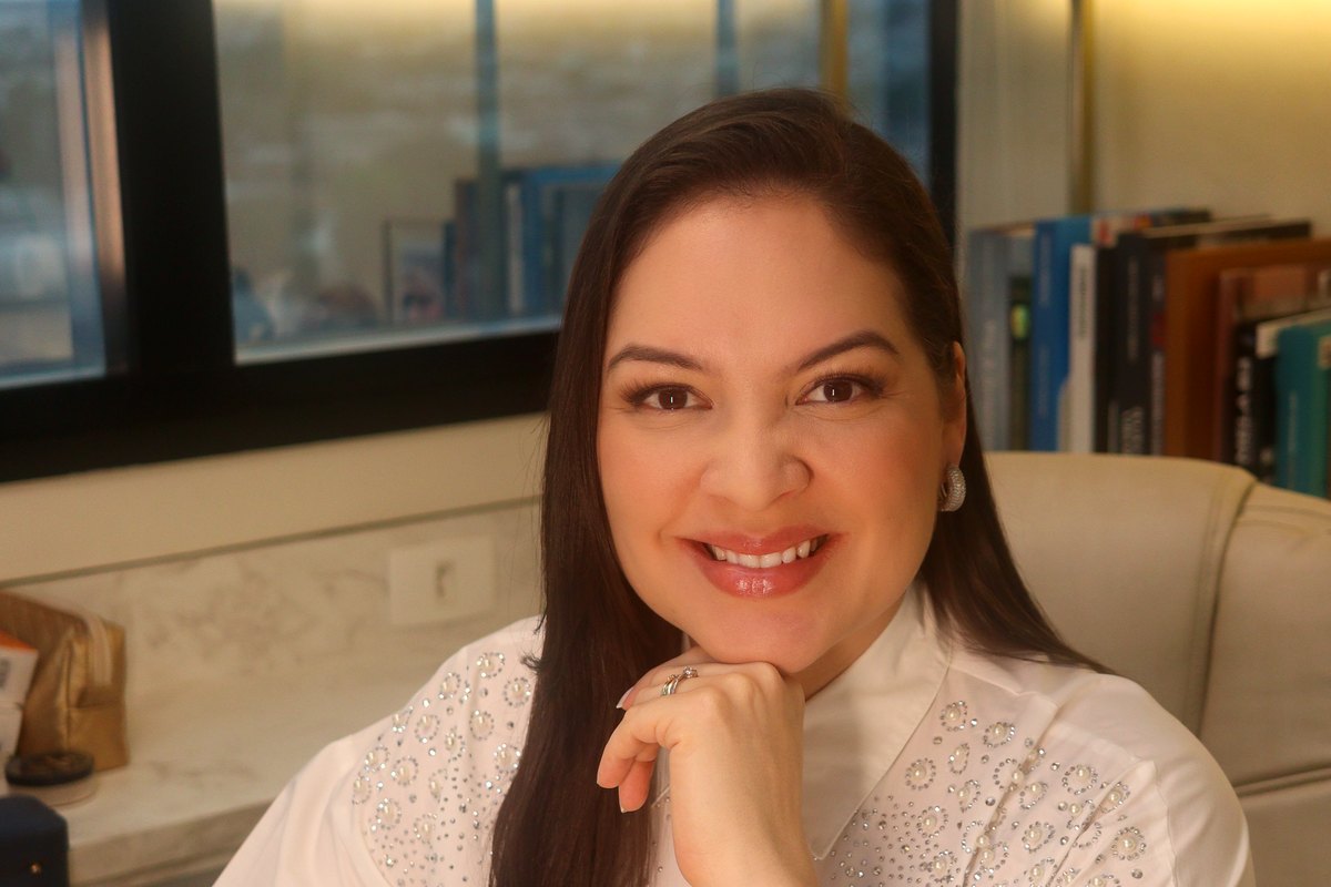 Dra. Aline Záu sentada à mesa de atendimento, com a janela do 16º andar ao fundo.